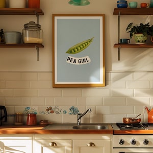 Può includere: Una cucina retrò con frigorifero azzurro, mobili bianchi e una stufa vintage. Una stampa incorniciata con un'illustrazione di baccello di pisello e la scritta "PEA GIRL" è appesa al muro. Mensole con vari utensili da cucina.