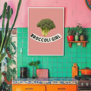 Könnte beinhalten: Buntes Kücheninterieur mit rosa Wand und grün gefliestem Fliesenspiegel. Ein gerahmter Druck mit einem Brokkoli-Bild und dem Text "BROCCOLI GIRL" hängt über orangefarbenen Schränken. Pflanzen und Küchenutensilien ergänzen die Szene.