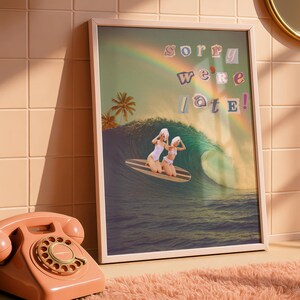 May include: A framed art print with a vintage aesthetic. It depicts two women on a surfboard, riding a wave, with a rainbow and palm trees. The text "Sorry we're late!" is displayed. The color scheme includes blues, greens, and pinks, evoking a summery vibe.