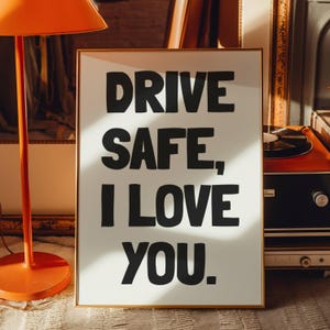 May include: A framed print with the words "DRIVE SAFE, I LOVE YOU" in bold black letters on a white background. The frame is gold-colored. An orange lamp and vintage record player are in the background.