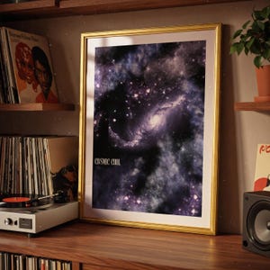 May include: A framed print with a gold border displays a cosmic galaxy scene in shades of purple, black, and white. The text "COSMIC COOL" is printed in white at the bottom. The print is displayed on a wooden shelf with other items.