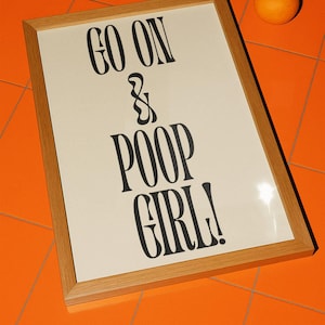 Puede incluir: Un cartel enmarcado con el texto "GO ON & POOP GIRL!" en negro y en negrita. El cartel tiene un fondo crema y un marco de madera marrón claro. Una naranja está sobre una superficie de baldosas naranjas.