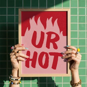 Puede incluir: Un estampado enmarcado con las palabras "UR HOT" en rojo, sobre un fondo rosa con un diseño de llamas rojas. El marco es de color marrón claro. La imagen está sostenida por manos adornadas con anillos y pulseras.