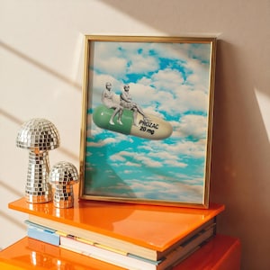 Könnte beinhalten: Ein gerahmter Kunstdruck mit goldenem Rahmen zeigt ein surreales Bild von zwei Personen, die auf einer großen grünen und weißen Prozac-Pille sitzen, die in einem blauen Himmel mit Wolken schwebt. Der Text "PROZAC 20 mg" ist auf der Pille zu sehen. Zwei verspiegelte pilzförmige Deko-Objekte stehen auf einem orangefarbenen Regal.