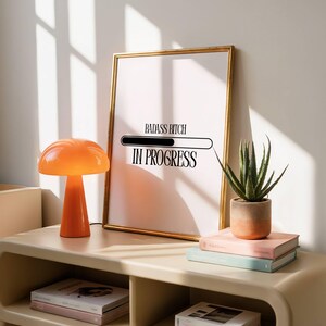 Può includere: Una stampa incorniciata con la scritta "BADASS BITCH IN PROGRESS" in nero. A sinistra c'è una lampada arancione a forma di fungo. Una piccola pianta grassa in vaso e libri sono su una mensola beige.