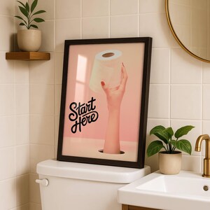 以下が含まれることがあります： ピンク色の背景にトイレットペーパーのロールを持っている手を描いた額入りのアートプリント。 黒いテキスト「Start Here」はスタイリッシュなフォントで表示されます。 アートワークはトイレの上に表示されます。