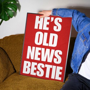 Pode incluir: Uma placa retangular vermelha com as palavras "HE'S OLD NEWS BESTIE" em letras maiúsculas brancas. A placa é segurada por uma pessoa vestindo uma jaqueta jeans. A placa está apoiada em um sofá dourado.