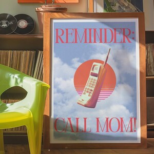 Pode incluir: Pôster emoldurado com um design retrô apresentando uma ilustração de telefone celular vintage contra um céu nublado e um sol poente. O texto diz "REMINDER: CALL MOM!" em uma fonte rosa arrojada.