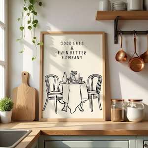 May include: A framed print with the text "GOOD EATS & EVEN BETTER COMPANY" above a black and white illustration of a table set for two. The image is in a kitchen setting with copper pots, jars, and a wooden cutting board.