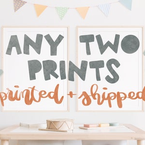 May include: Two framed prints with the words "ANY TWO PRINTS printed + shipped" in gray and orange lettering. The prints are displayed on a white wall with colorful bunting and a small table with toys.