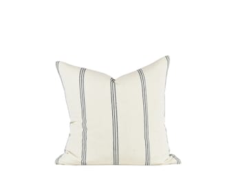 May include: A square decorative pillow with a cream-colored background and vertical navy blue stripes. The pillow has a soft, textured appearance, suitable for home decor.