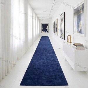 Puede incluir: Una alfombra pasillera larga y rectangular de color azul marino sólido y profundo con una textura de pelo corto y denso. La alfombra está colocada centralmente en el suelo de un pasillo blanco brillante, extendiéndose desde el frente al fondo.