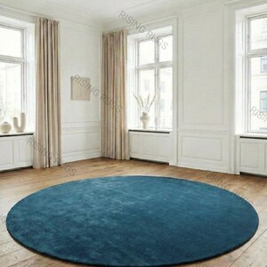 May include: A large, round teal rug dominates a room with hardwood floors. The rug is centered in the room, surrounded by white walls, windows, and beige curtains. The room is well-lit, creating a bright and airy atmosphere. The rug's texture is visible.