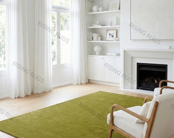 May include: A modern living room featuring a large, rectangular olive green rug. The rug is placed on a light wood floor, with white curtains and a fireplace in the background. Two white armchairs with wooden frames are also visible.