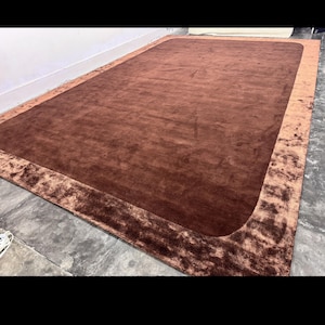May include: A large, rectangular brown rug with a darker brown border. The rug has a plush texture and is placed on a concrete floor. The border has a slightly raised, textured appearance.