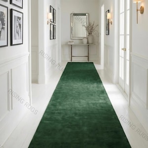 Puede incluir: Una larga alfombra verde esmeralda en un pasillo con paredes y molduras blancas. La alfombra se extiende por el centro del pasillo, creando un camino visual. Fotos enmarcadas en negro y apliques adornan las paredes.