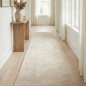 May include: A neutral-toned, rectangular area rug in a hallway setting. The rug is a light beige color and has a soft, textured appearance. The hallway features light wood flooring, white walls, and natural light from a window.