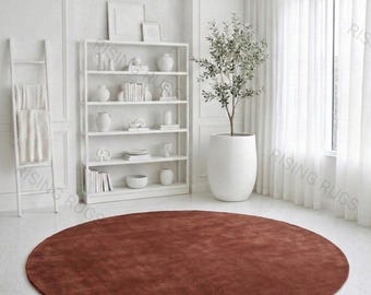 Puede incluir: Una alfombra circular grande de color terracota sólido con una textura suave y uniforme. La alfombra está colocada en el centro sobre un suelo de baldosas blancas en una habitación luminosa, creando un contraste con la decoración blanca.