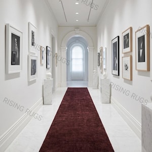 Puede incluir: Una alfombra de pasillo larga y rectangular de color burdeos intenso con una superficie de pelo texturizado. La alfombra está colocada en el centro sobre un suelo de mármol blanco en un pasillo luminoso, extendiéndose hacia un arco.