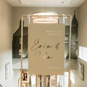 May include: A beige wedding sign with gold lettering that reads "Here's to forever Erin & John 28|06|24".