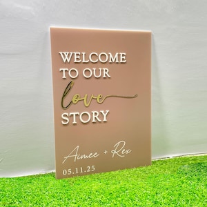 May include: A rectangular sign with the words "WELCOME TO OUR love STORY" in white and gold lettering. The names "Aimee + Rex" and the date "05.11.25" are also displayed in white. The sign is a muted pink color.