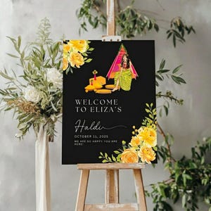 May include: A black sign on an easel with the text "WELCOME TO ELIZA'S Haldi OCTOBER 11, 2025 WE ARE SO HAPPY YOU ARE HERE!". The sign features yellow floral accents and an illustration of a woman in traditional attire.