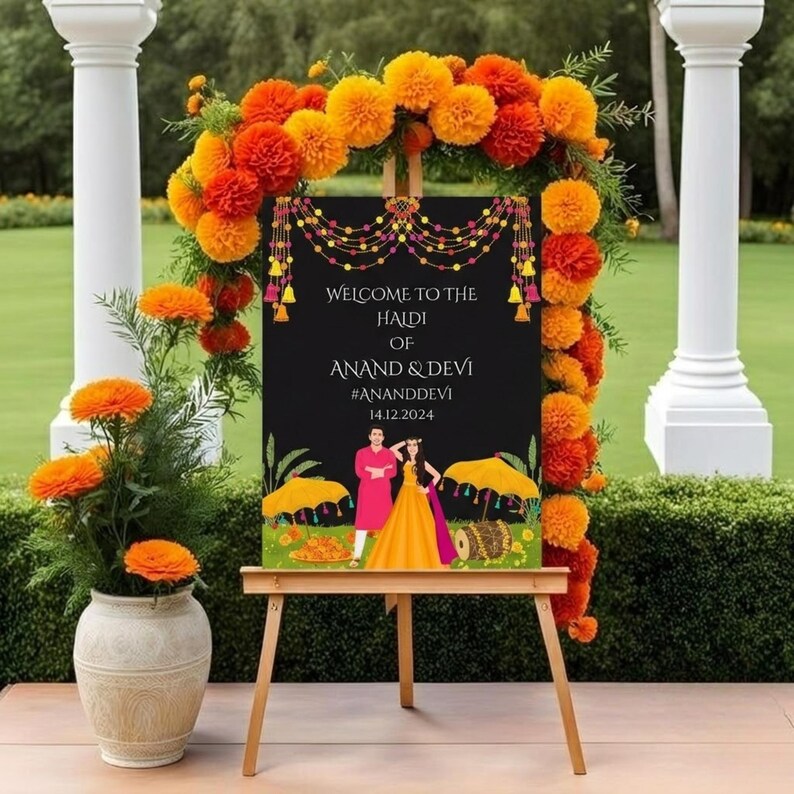 May include: A wooden easel displays a black sign with the text "WELCOME TO THE HALDI OF ANAND & DEVI #ANANDDEVI 14.12.2024". The sign is decorated with a floral garland of orange and yellow flowers. A vase of orange flowers is on the left.