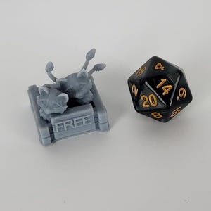 Könnte beinhalten: Zwei graue, 3D-gedruckte Kätzchen in einer grauen Box mit dem Wort "FREE" auf der Vorderseite. Ein schwarzer 20-seitiger Würfel mit goldenen Zahlen befindet sich im Hintergrund.
