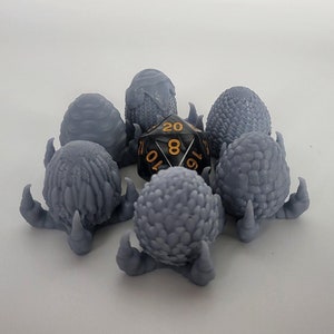 Puede incluir: Seis figuritas grises en forma de huevo de dragón impresas en 3D con un dado negro de 20 caras en el centro.