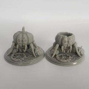 Puede incluir: Dos figuras grises con forma de calabaza y patas de araña. Una tiene la parte superior sólida, la otra es hueca. Ambas se asientan sobre una base circular con elementos decorativos. Para decoración de Halloween o fantasía.