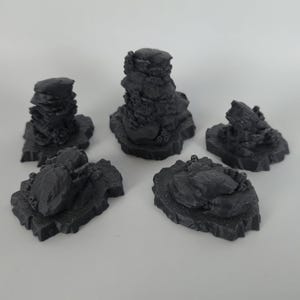 Pode incluir: Cinco formações rochosas em miniatura cinza escuro de vários tamanhos e formas. As rochas têm uma superfície áspera e texturizada e estão dispostas sobre um fundo branco. Poderiam ser usadas para um comboio modelo ou um jogo de tabuleiro.