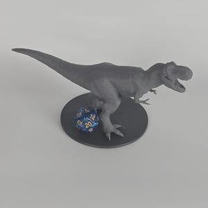 May include: A gray Tyrannosaurus Rex dinosaur figurine standing on a circular base. Next to the dinosaur's feet is a blue twenty-sided die with gold numbers, featuring the number 20 clearly visible on the front face of the gaming die.
