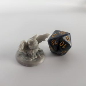 K&ouml;nnte beinhalten: Eine graue, 3D-gedruckte Miniatur eines gefl&uuml;gelten Wesens mit einem runden Sockel. Das Wesen hat einen pelzigen K&ouml;rper und gro&szlig;e Fl&uuml;gel. Ein schwarzer und goldener 12-seitiger W&uuml;rfel befindet sich neben der Miniatur.