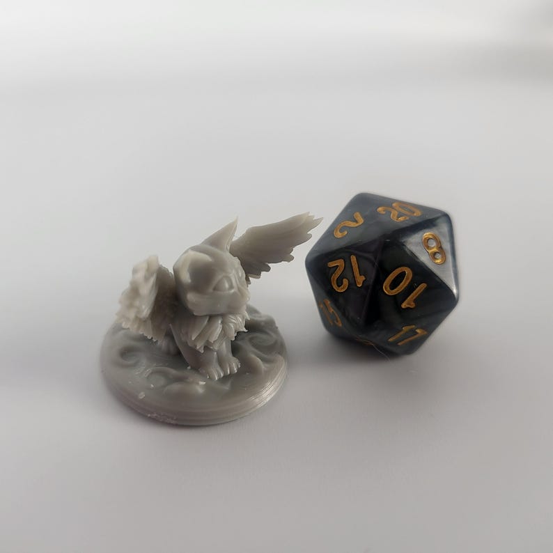 K&ouml;nnte beinhalten: Eine graue, 3D-gedruckte Miniatur eines gefl&uuml;gelten Wesens mit einem runden Sockel. Das Wesen hat einen pelzigen K&ouml;rper und gro&szlig;e Fl&uuml;gel. Ein schwarzer und goldener 12-seitiger W&uuml;rfel befindet sich neben der Miniatur.