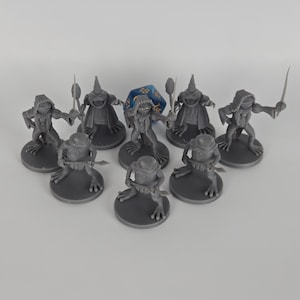 May include: A collection of gray miniature frog figurines, each posed with a weapon or staff, arranged on circular bases. A blue, multi-sided die is placed among the figurines. The figurines are detailed and appear to be part of a tabletop game.