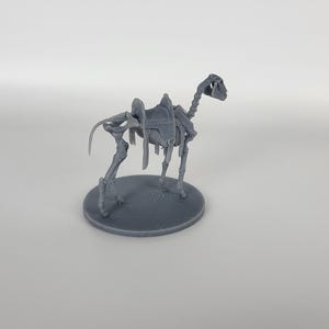 May include: A gray plastic miniature of a skeletal camel with a saddle on its back. The camel is standing on a round base.