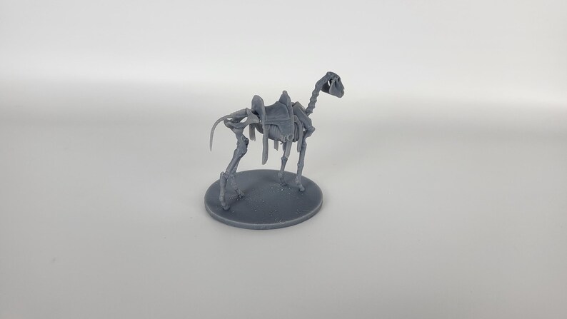 May include: A gray plastic miniature of a skeletal camel with a saddle on its back. The camel is standing on a round base.