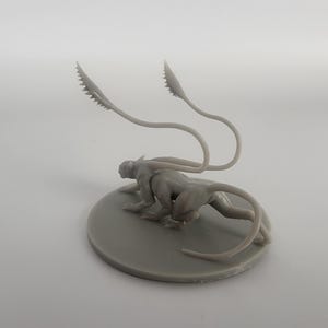 以下が含まれることがあります： 2本の細長い尻尾を持つ生き物のグレーの3Dプリントミニチュア。生き物は丸い土台の上にしゃがんでいます