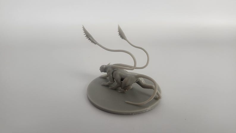 以下が含まれることがあります： 2本の細長い尻尾を持つ生き物のグレーの3Dプリントミニチュア。生き物は丸い土台の上にしゃがんでいます