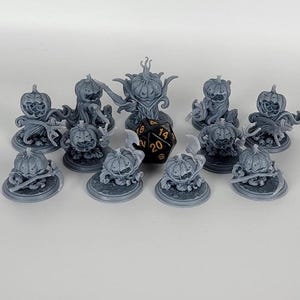 Può includere: Un set di 10 figurine di zucca dipinte di grigio, ognuna con un design unico e un dado a 20 facce nero e oro al centro.