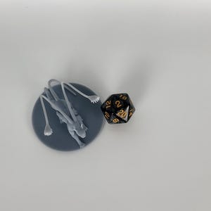 以下が含まれることがあります： 灰色の3Dプリントされたミニチュアは、長い脚と尾を持つ生き物を表しており、灰色のベースの上に立っています。ミニチュアの隣には、金色の数字が書かれた黒い20面ダイスがあります。