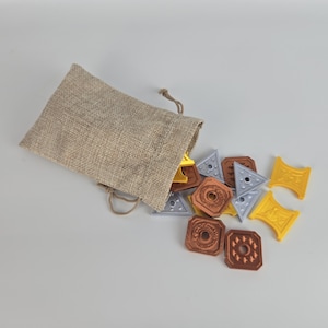 May include: A beige burlap drawstring bag with assorted game pieces spilling out. The game pieces are shaped as triangles, squares, and rectangles, in shades of yellow, silver, and copper.