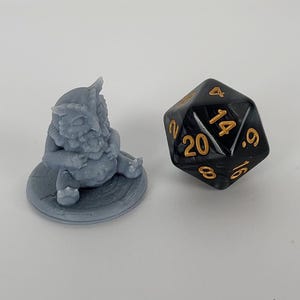 May include: A small gray owl-like creature figurine sitting on a round base. The figurine is holding a small object in its hands. A black 20-sided die with gold numbers is in the background.