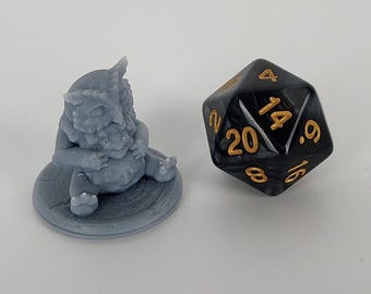 May include: A small gray owl-like creature figurine sitting on a round base. The figurine is holding a small object in its hands. A black 20-sided die with gold numbers is in the background.