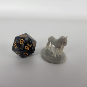 May include: A black and silver 20-sided die with gold numbers sits next to a gray miniature horse figurine on a gray base.