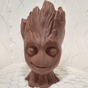Puede incluir: Un macetero de cerámica marrón con forma de la cabeza del personaje Groot de la franquicia de películas Guardianes de la Galaxia. El macetero tiene una cara sonriente y una superficie texturizada.