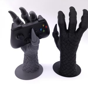 May include: Two matte black claw-shaped stands with textured, scale-like surfaces and circular bases. One stand holds a black wireless video game controller with multi-coloured action buttons, while the other stand is empty and upright.