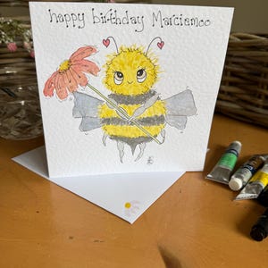 Puede incluir: Tarjeta de cumpleaños hecha a mano con una ilustración en acuarela de una abeja de dibujos animados sosteniendo una flor. La abeja es amarilla y negra con alas grises. La tarjeta dice "happy birthday Marciamoo" en tinta negra.