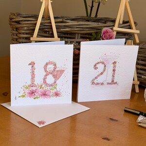 May include: Two white greeting cards with watercolor illustrations. One card displays the number "18" adorned with pink flowers and a cocktail glass. The other card shows the number "21" with similar floral accents and a cocktail glass.