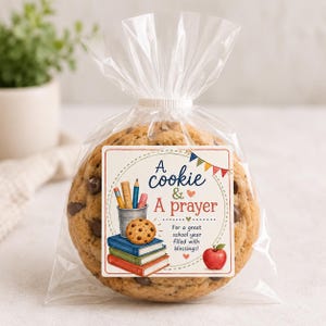 Könnte beinhalten: Ein großer Schokoladenkeks in einer durchsichtigen Plastiktüte mit einem quadratischen Etikett, das Bücher, Stifte, einen Apfel und den Text A cookie & A prayer For a great school year filled with blessings! in Blau und Rot zeigt.